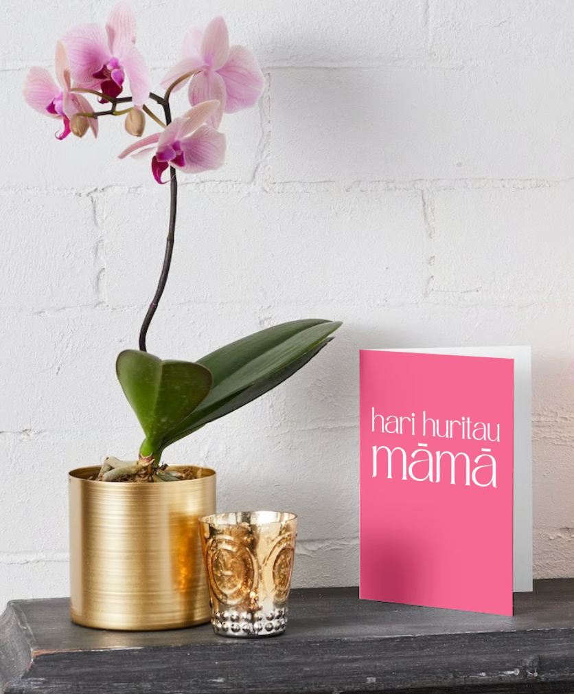 Te Reo Māori Card - reads Hari Huritau Māmā on the front cover. The cover is bright pink with contrasting white text.
The card is standing up on a bench in front of a white brick wall with a potted orchid and a candle next to it.